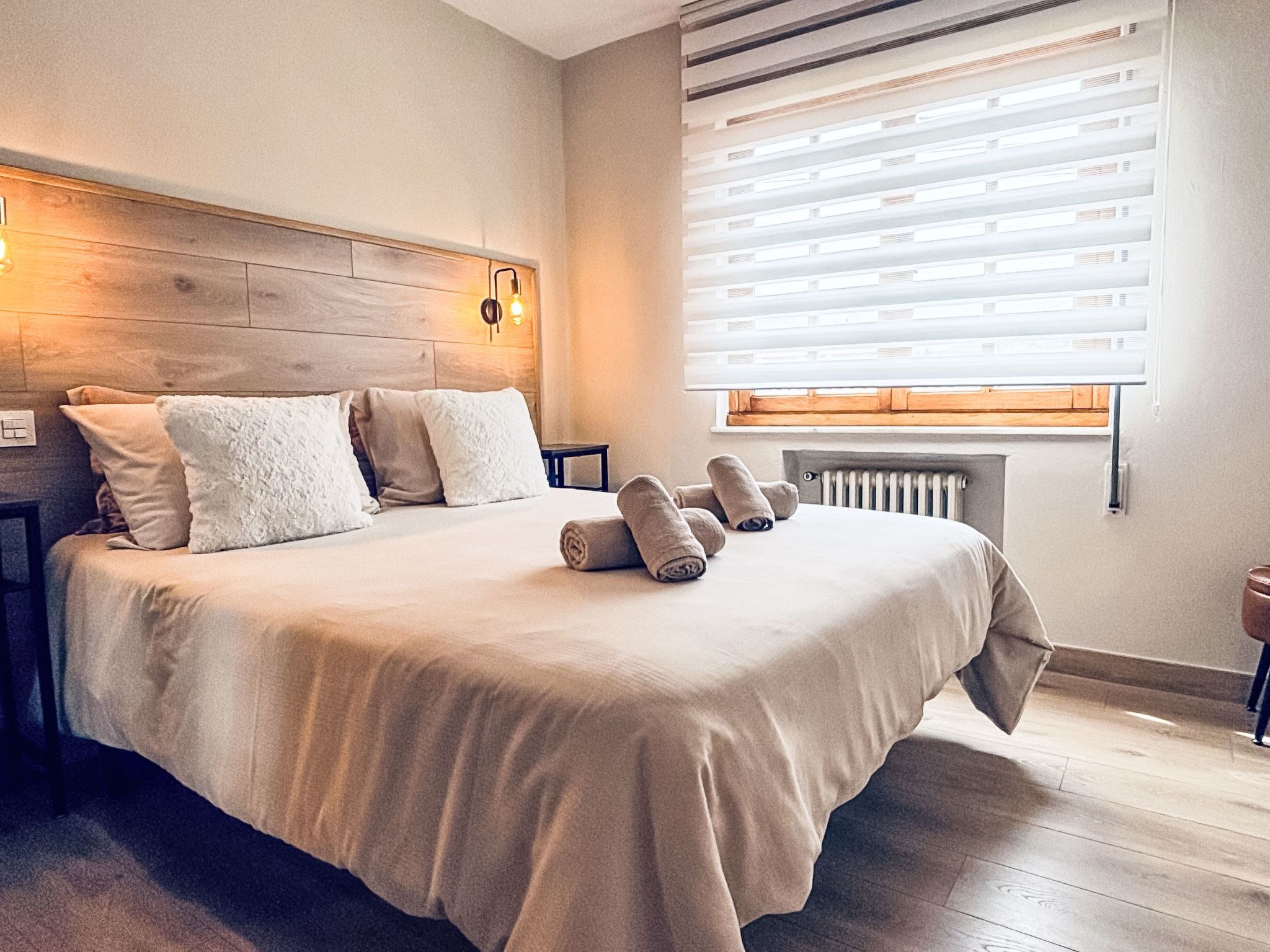 Una cama bien hecha con almohadas y toallas enrolladas, un cabecero de madera, una mesita de noche y un radiador.
