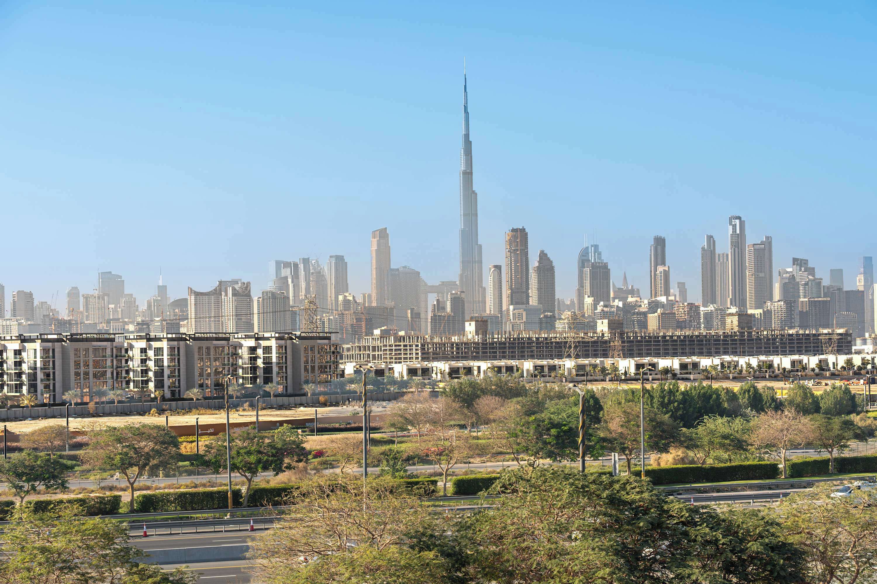 Panorama urbano con un grattacielo dominante, probabilmente un quartiere degli affari con edifici moderni, un'ampia area verde con alberi e cielo sereno.