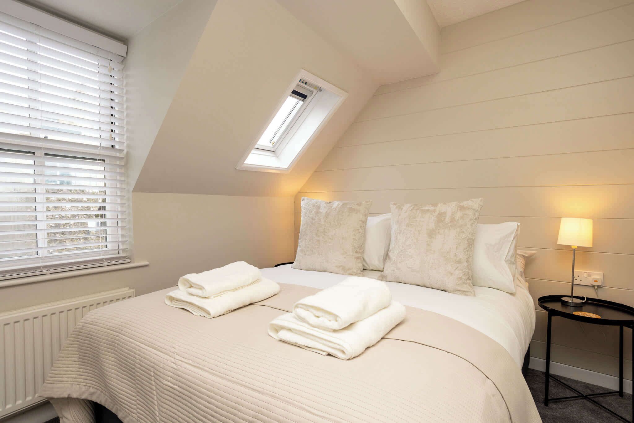 A bedroom with a sloped ceiling, a skylight, a bed with white bedding, a bedside table with a lamp, and a radiator.