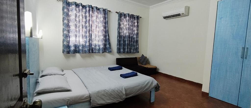 A single bed with pillows and a blanket, a small table with a chair, and a blue door.