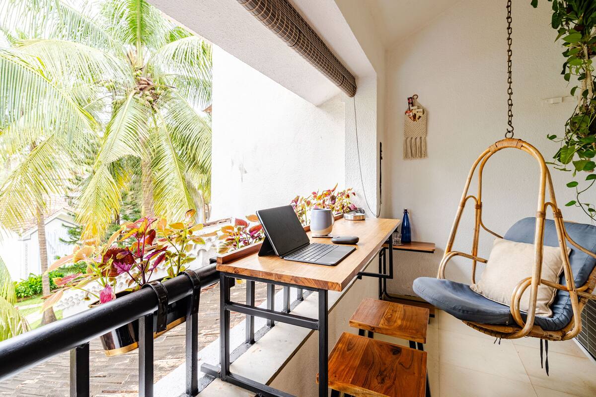 A room with a wooden desk, a laptop, a hanging chair, and a view of palm trees.