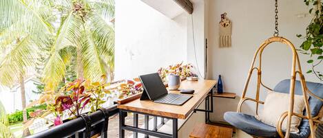A room with a wooden desk, a laptop, a hanging chair, and a view of palm trees.