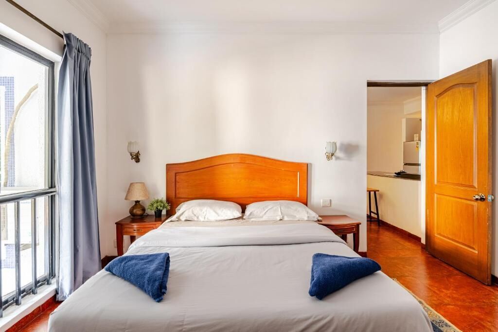 Quarto de hotel com uma cama grande, mesa de cabeceira com abajur, janela com cortinas e porta de madeira.