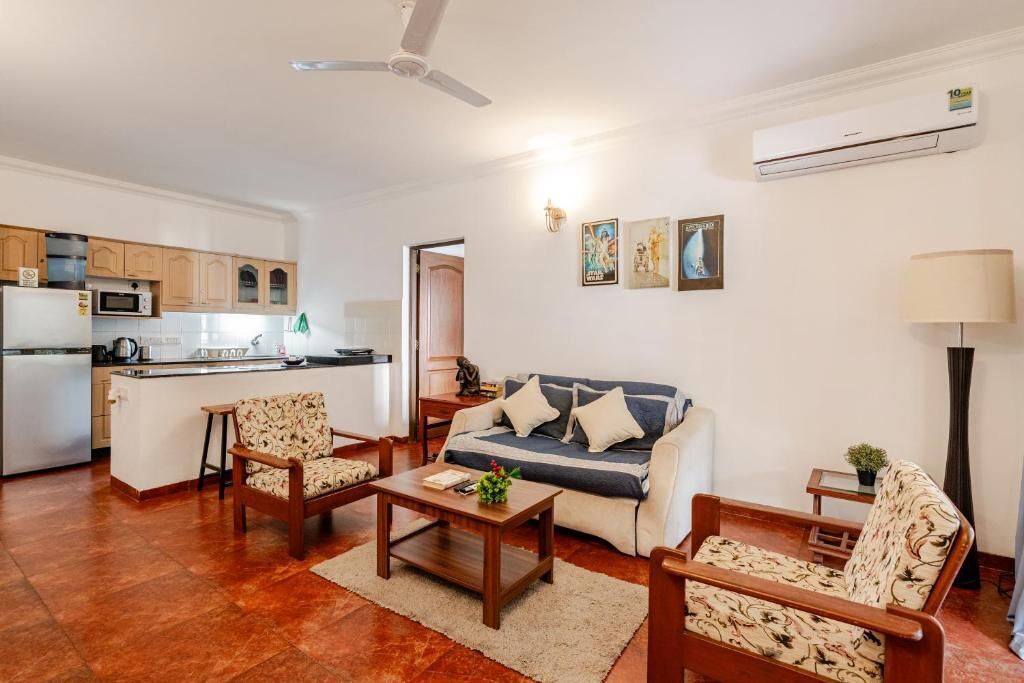 A living room with a sofa, two armchairs, a coffee table, and a lamp. There is a kitchen area with cabinets and appliances in the background.