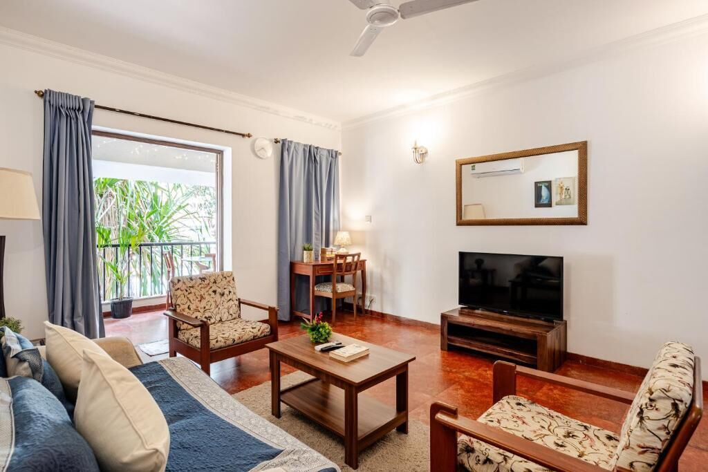 Uma sala de estar com sofá, poltronas, mesa de centro, televisão e varanda com cortinas.