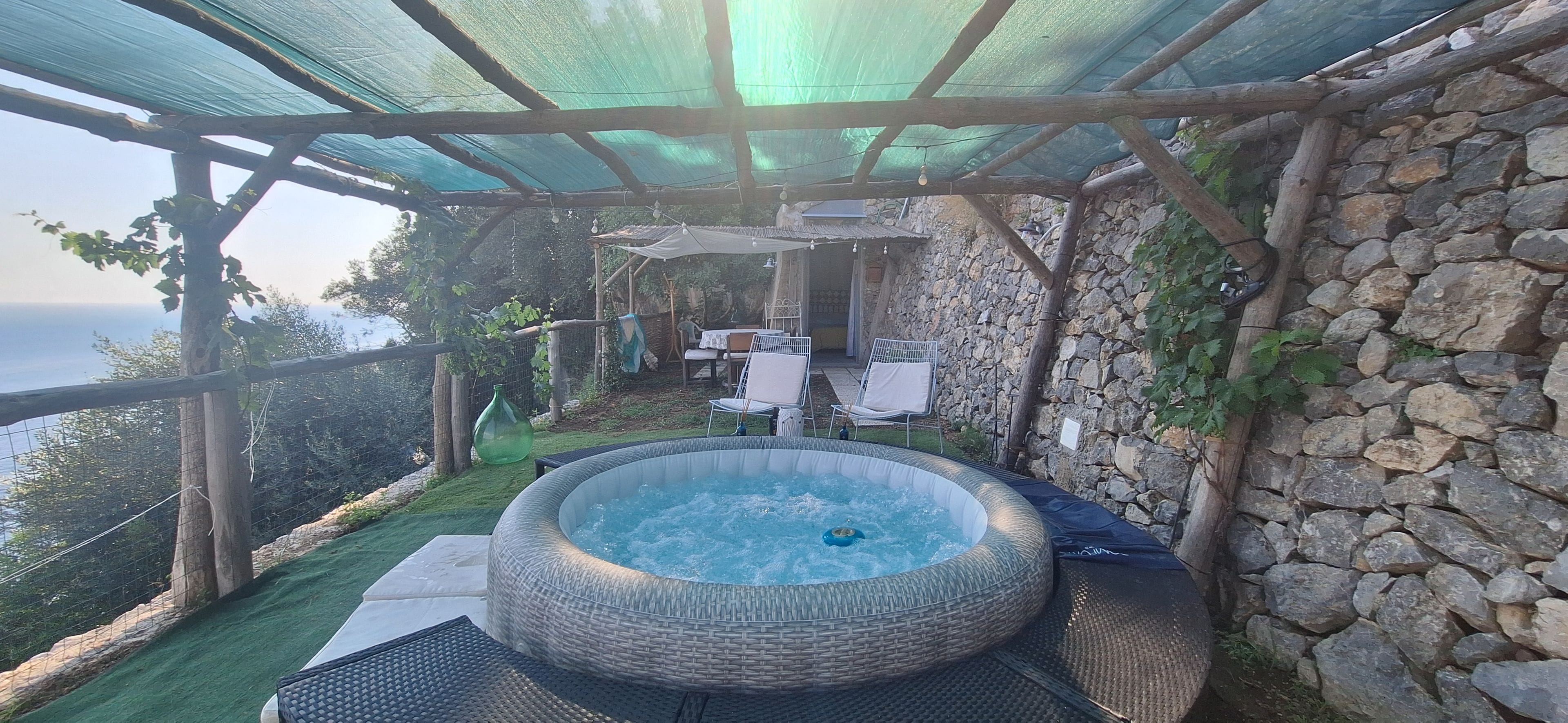A hot tub under a pergola with a stone wall and a view of the sea.
