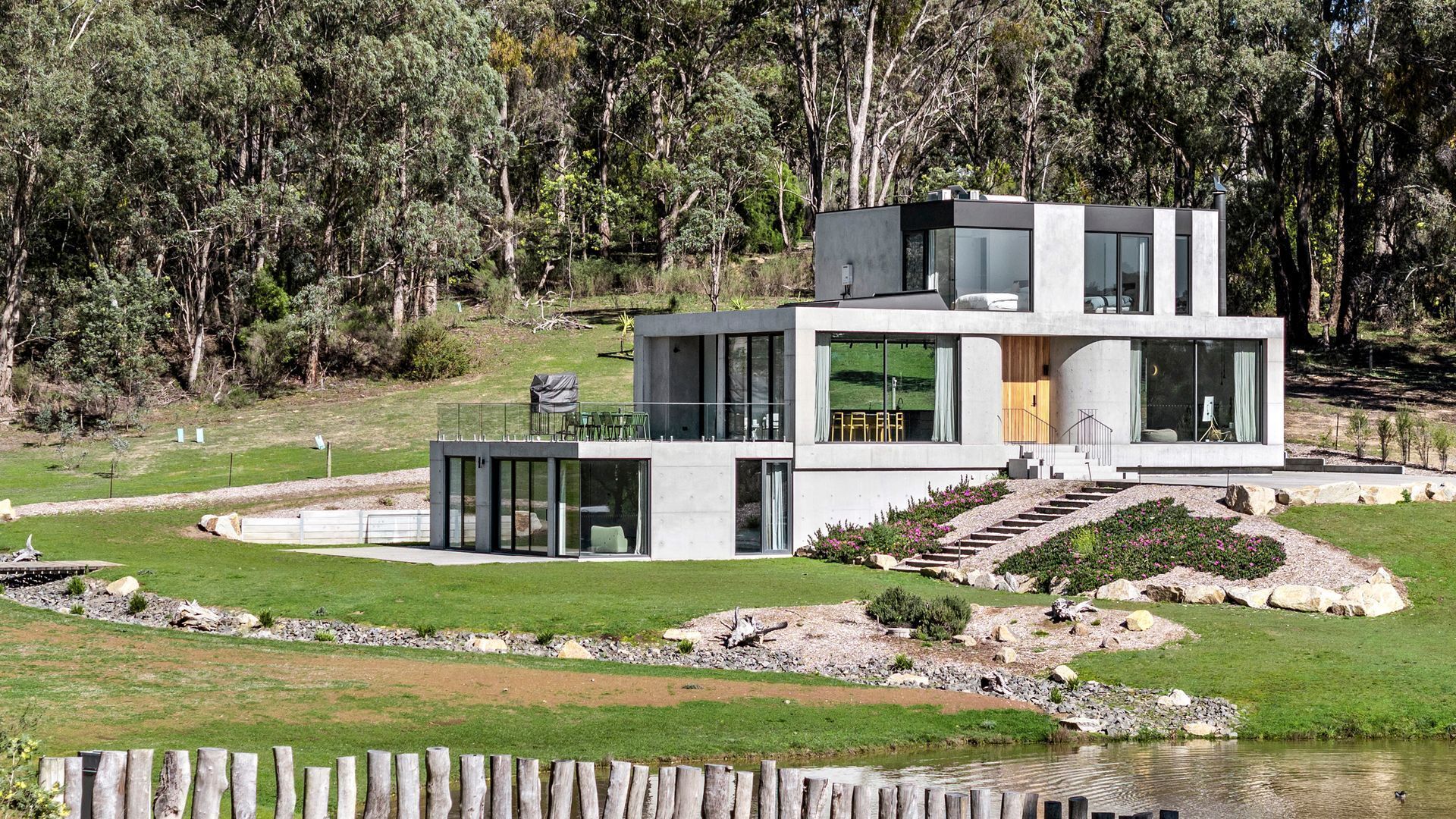 Une maison moderne à deux étages, dotée de grandes fenêtres en verre, d’une terrasse en bois et d’un chemin en pierre menant à l’entrée.