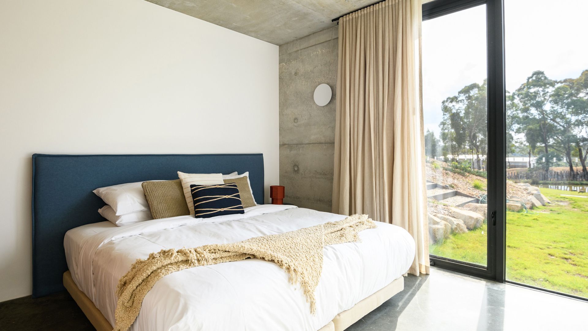 A modern bedroom with a large bed, a headboard, a bedside table, and a view of a garden through a large window.