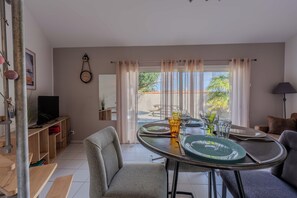 Un comedor con una mesa dispuesta para dos, un televisor sobre un mueble bajo y vistas al exterior a través de una ventana con cortinas traslúcidas.
