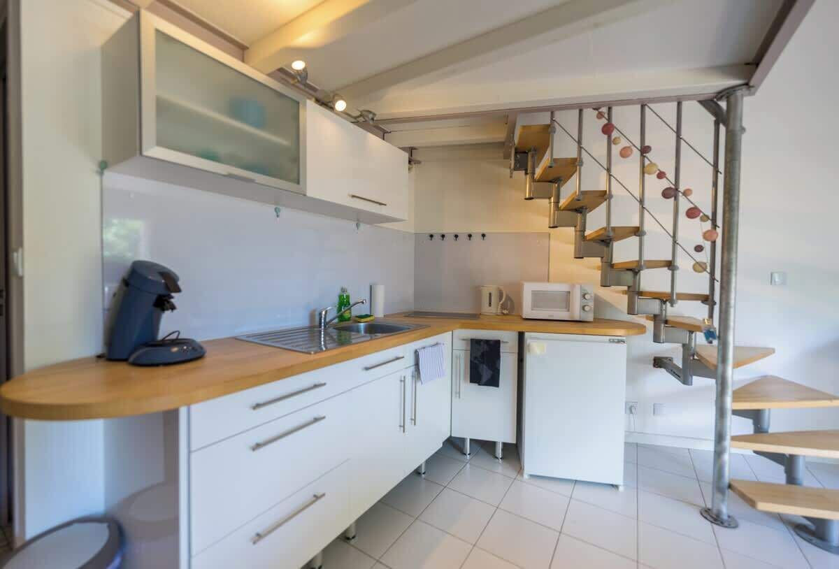 Una cucina compatta con mobili bianchi, un piano di lavoro in legno, un forno a microonde integrato e un piccolo frigorifero. Una scala con ringhiera in metallo conduce a una zona pranzo.
