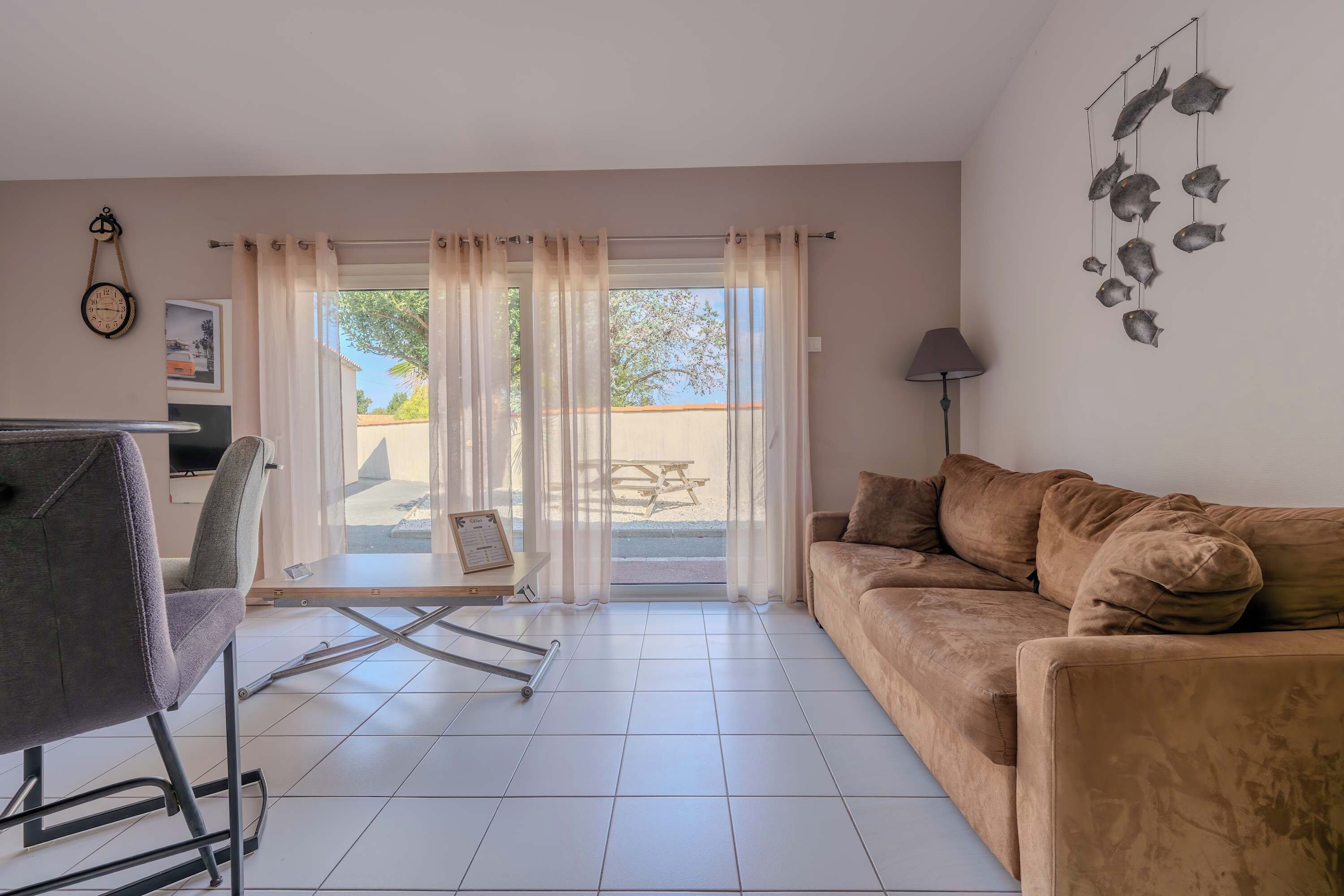 A living room with a sofa, a table, a chair, and a view through a glass door to an outdoor area.