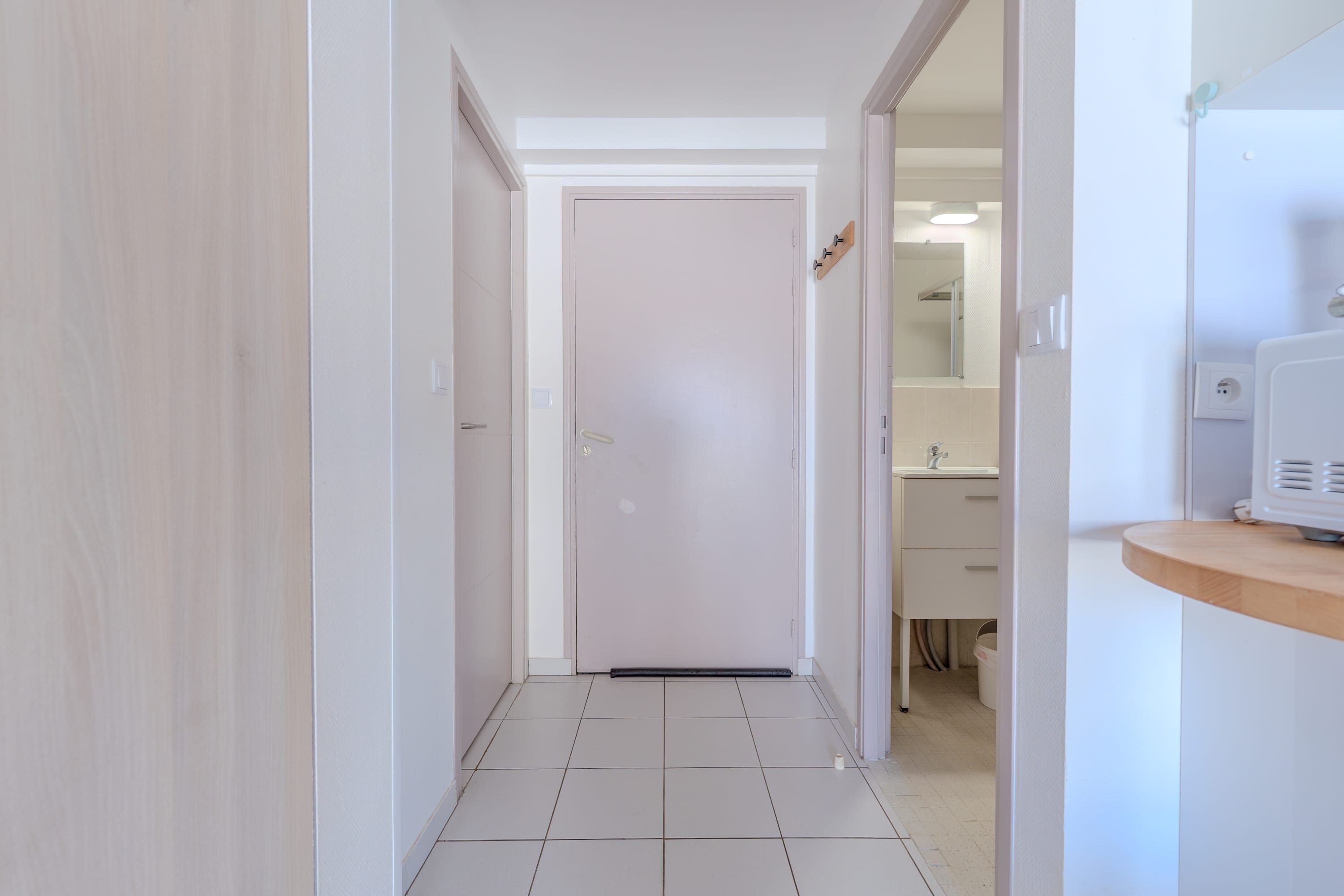 A hallway with a white door at the end, leading to a bathroom with a sink and mirror.