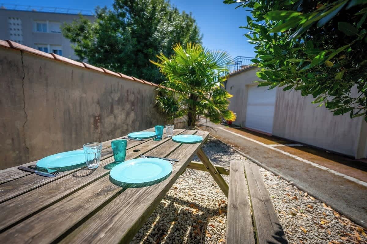 A wooden table set outdoors with plates and glasses, surrounded by gravel and a concrete wall.