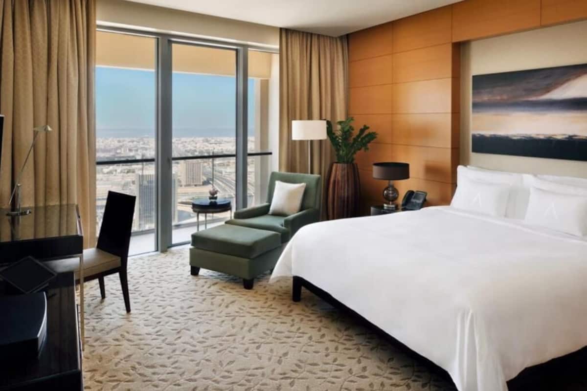 Habitación de hotel con una cama grande, un escritorio, una silla, una mesita y vistas a la ciudad a través de amplias ventanas.