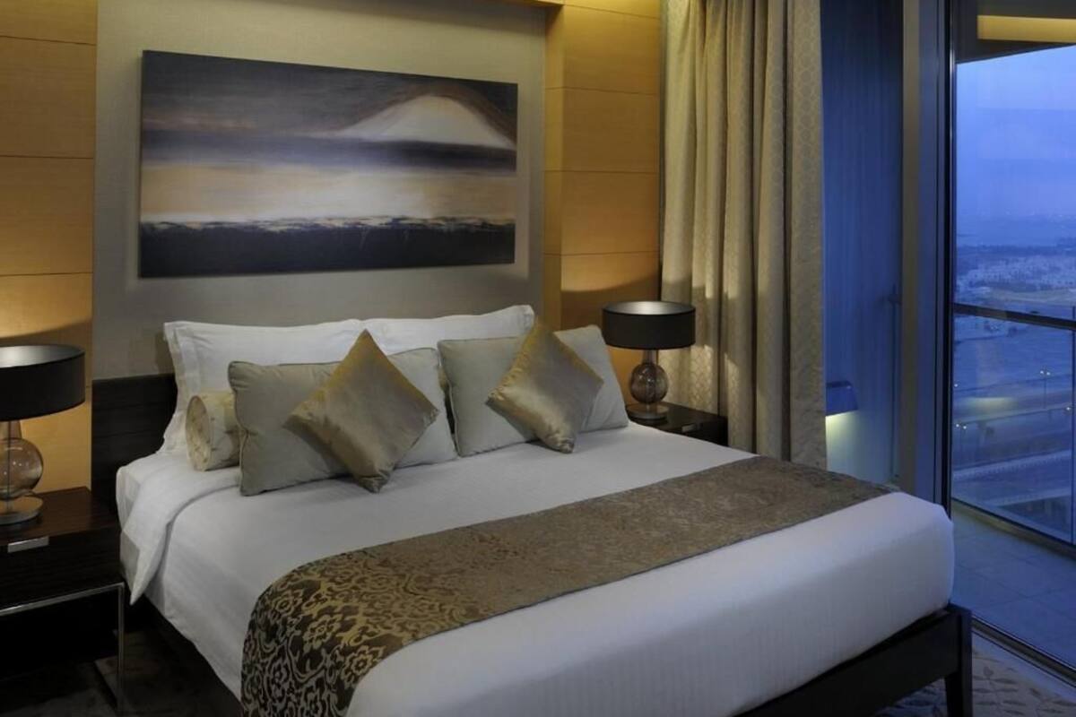 Habitación de hotel moderna con una cama grande, dos mesitas de noche con lámparas, un ventanal con vistas y un cuadro en la pared.