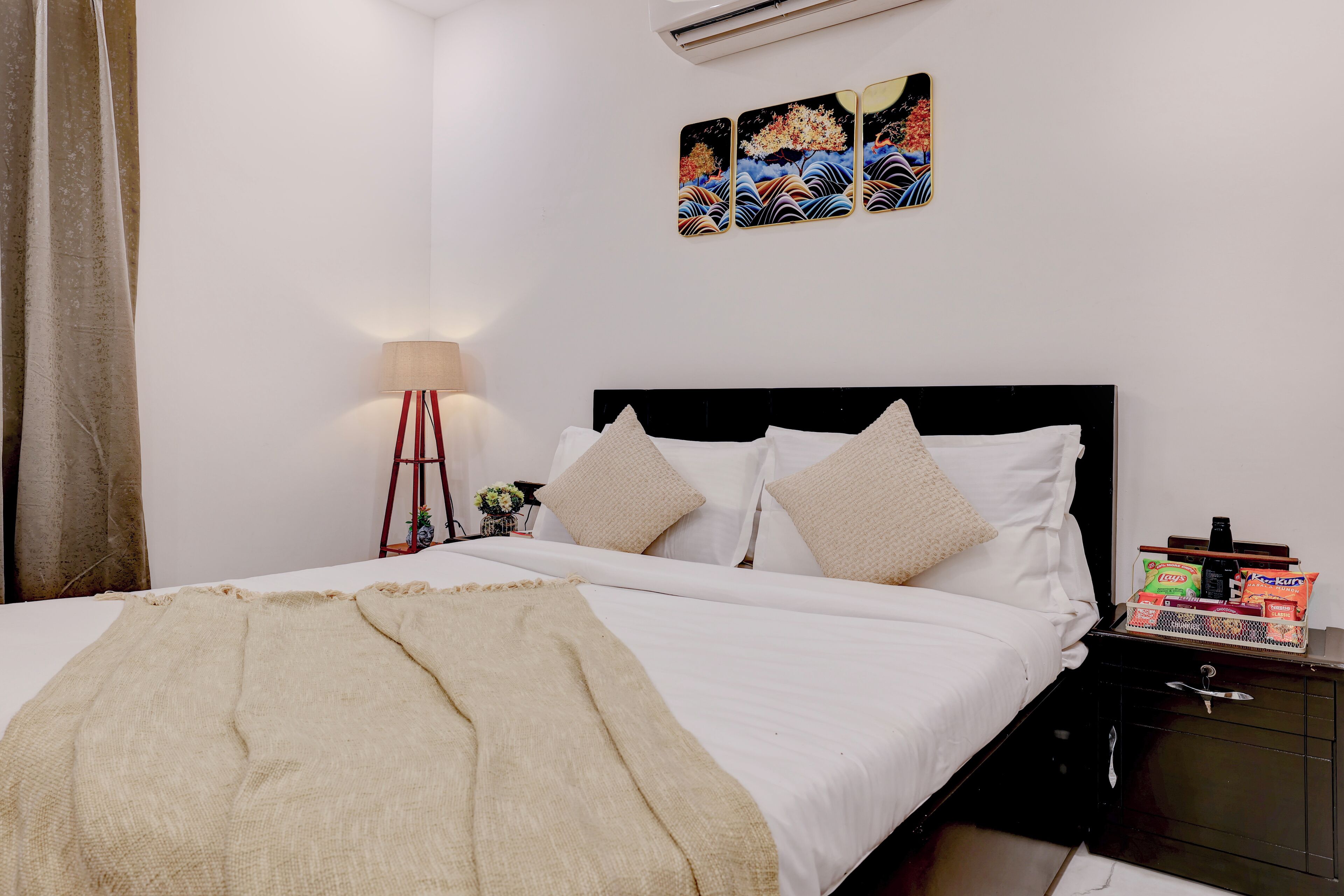 Una cama bien tendida con una manta color beige, una mesita de noche con aperitivos y una obra de arte colgada en la pared.
