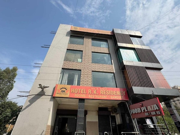 Un immeuble à plusieurs étages, sur lequel on peut lire l’enseigne « Hôtel R.K. Residency », indiquant la présence de chambres, d’un restaurant et de salles de réception.