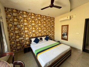 Une chambre avec un mur orné de motifs, un lit recouvert d’une courtepointe multicolore, un ventilateur de plafond, un appareil de climatisation et un miroir sur le mur.
