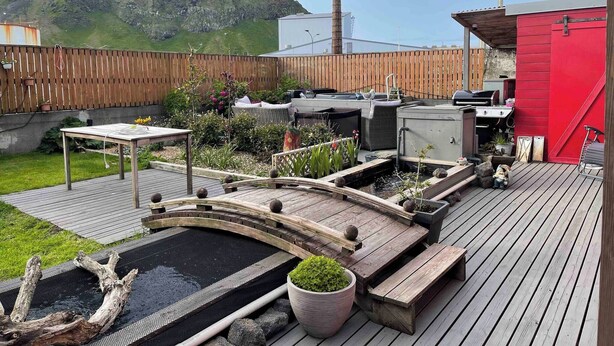 A deck with a small pond, wooden furniture, and a red building. - The Cozy Caravan Vestmannaeyjar (Vestmannaeyjar)