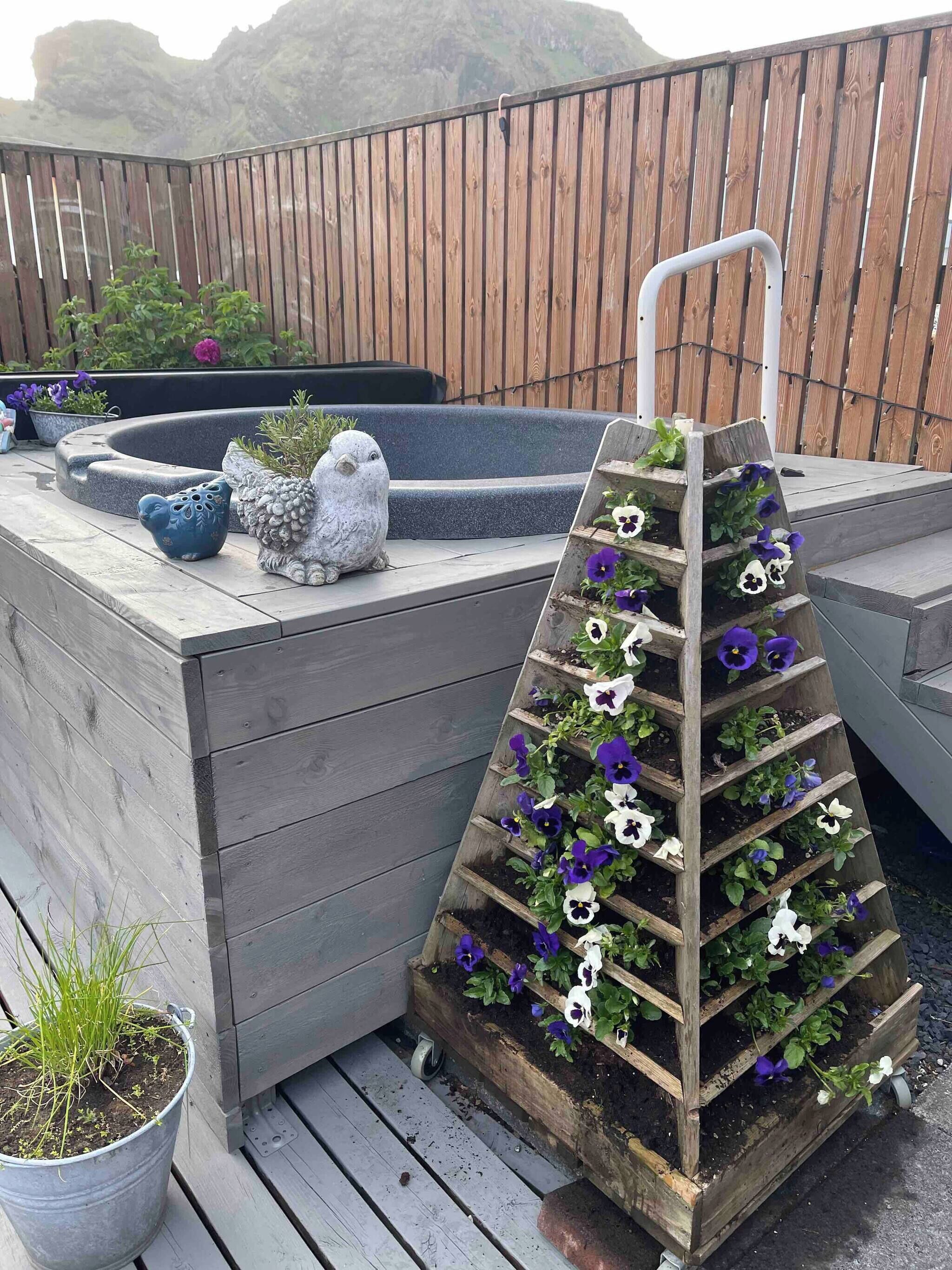 Una terraza de madera con un jardín vertical escalonado, lleno de flores, una escultura de pájaro y un pequeño estanque.