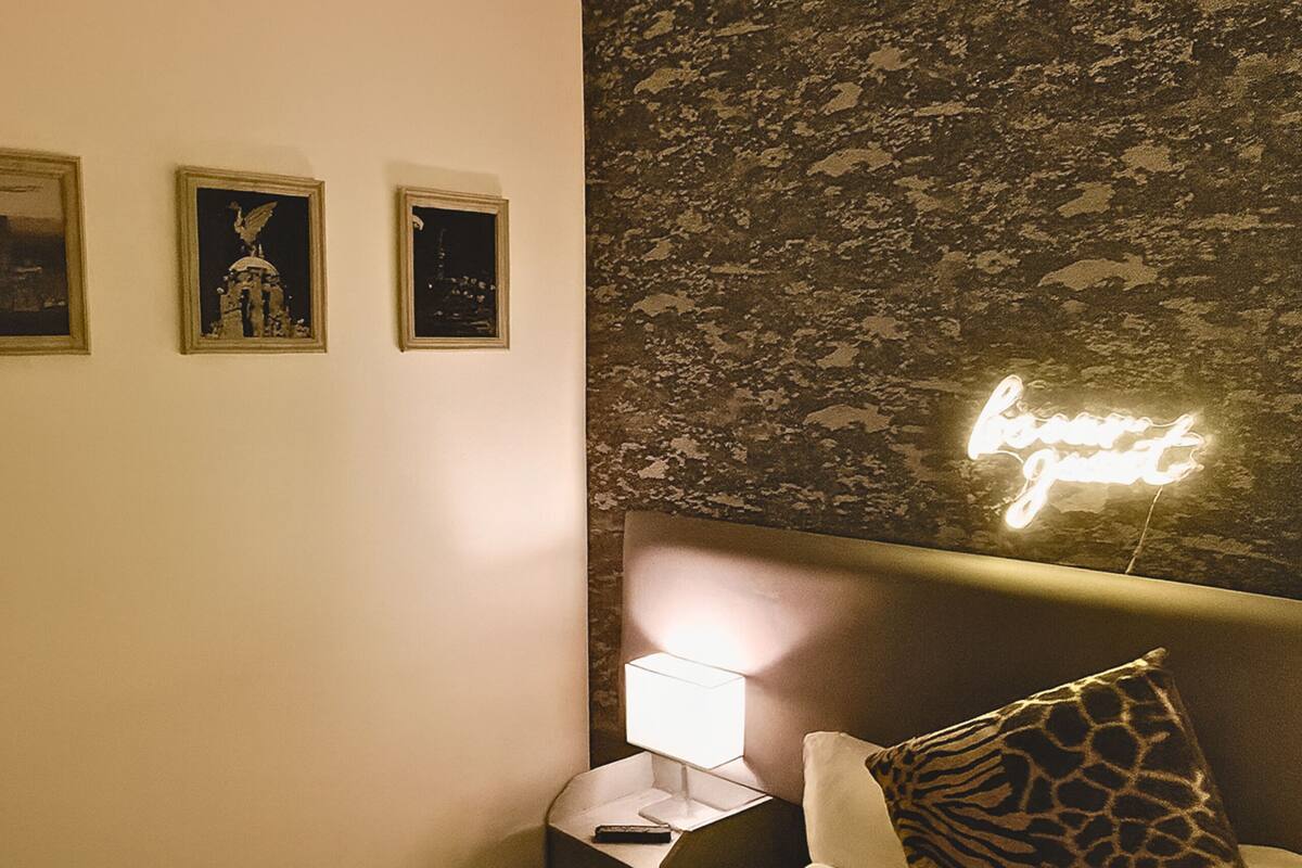 Une chambre à coucher avec une tête de lit, une table de chevet avec une lampe, une télécommande et trois tableaux encadrés accrochés au mur.