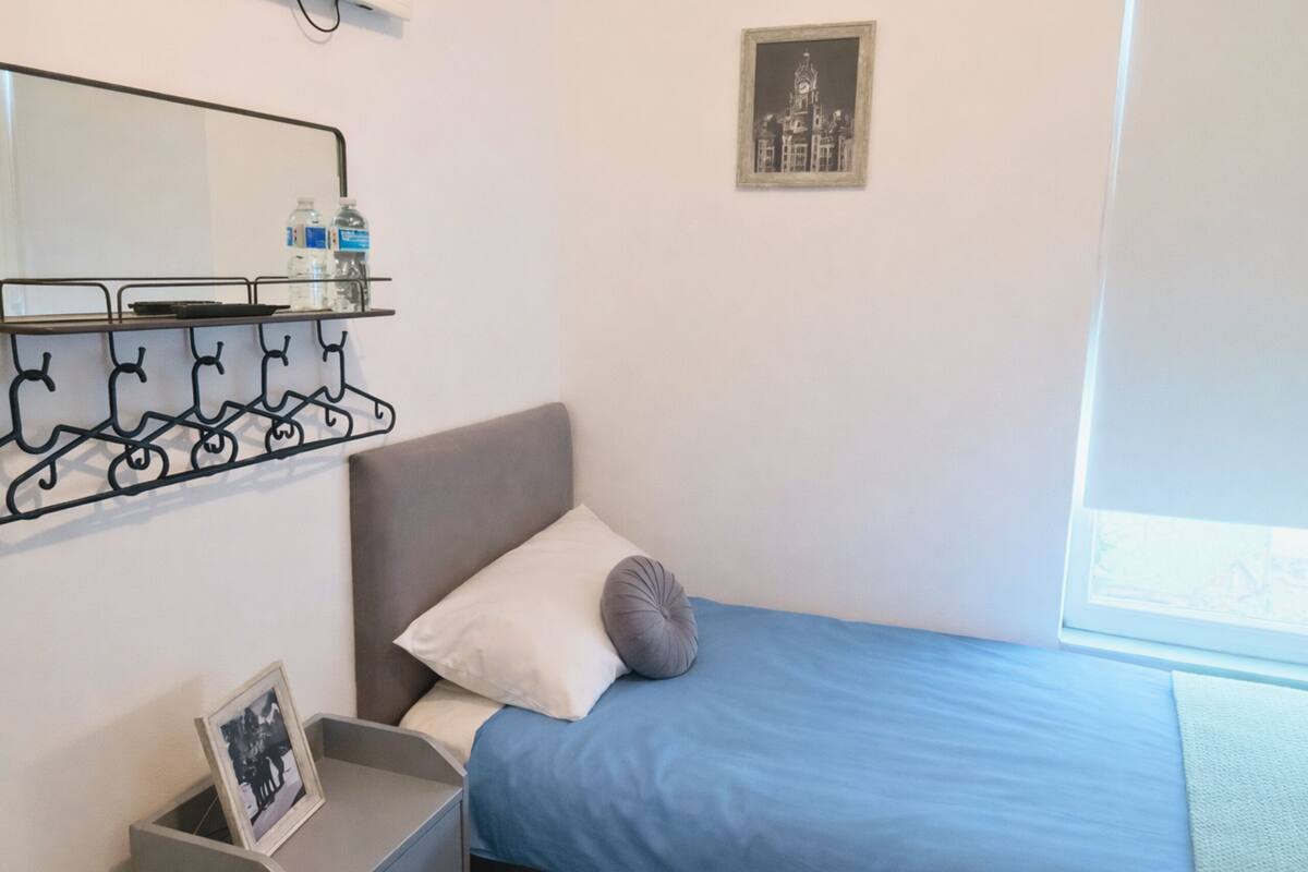 Une chambre à coucher avec un lit, une table de chevet, une étagère murale avec des bouteilles, un tableau encadré et une fenêtre.