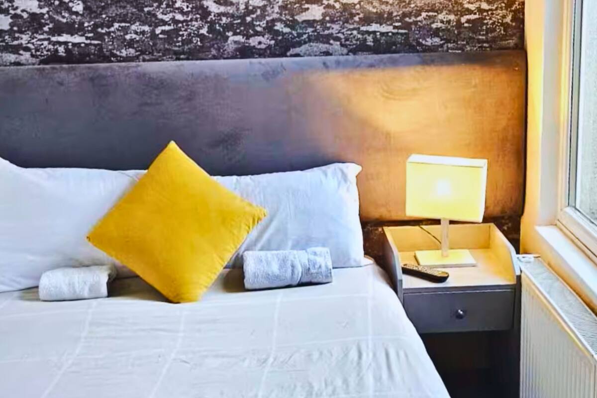 Un lit bien fait, orné d’oreillers blancs et jaunes, une table de chevet avec une lampe, et une fenêtre donnant sur l’extérieur.