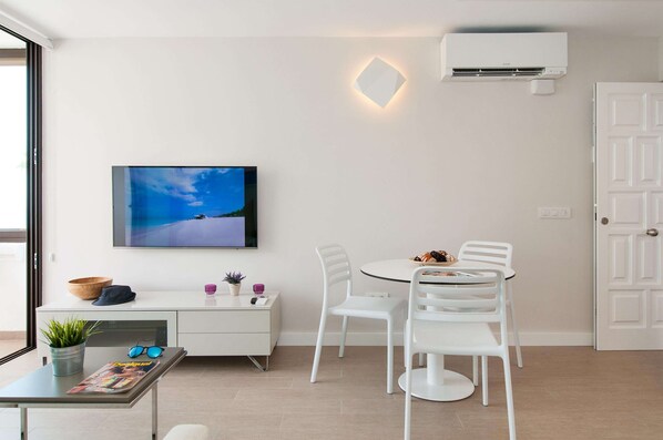 Uma sala de estar moderna com um sofá branco, uma mesa de jantar redonda com cadeiras brancas, uma TV de tela plana fixada na parede, um aparelho de ar condicionado branco e uma porta com luminária geométrica.