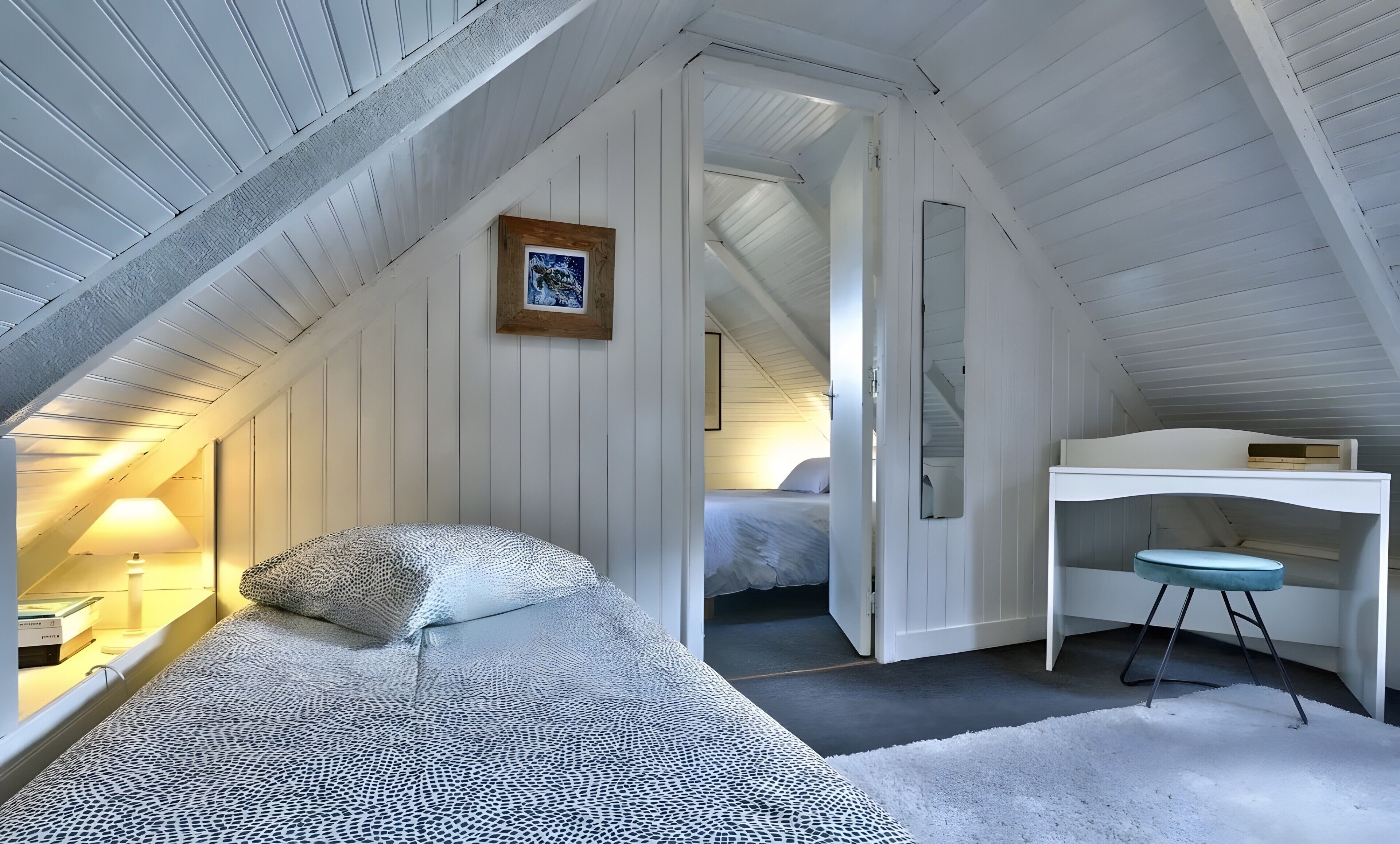 Un dormitorio con techo inclinado, cama con ropa de cama estampada, un escritorio con taburete y un espejo.