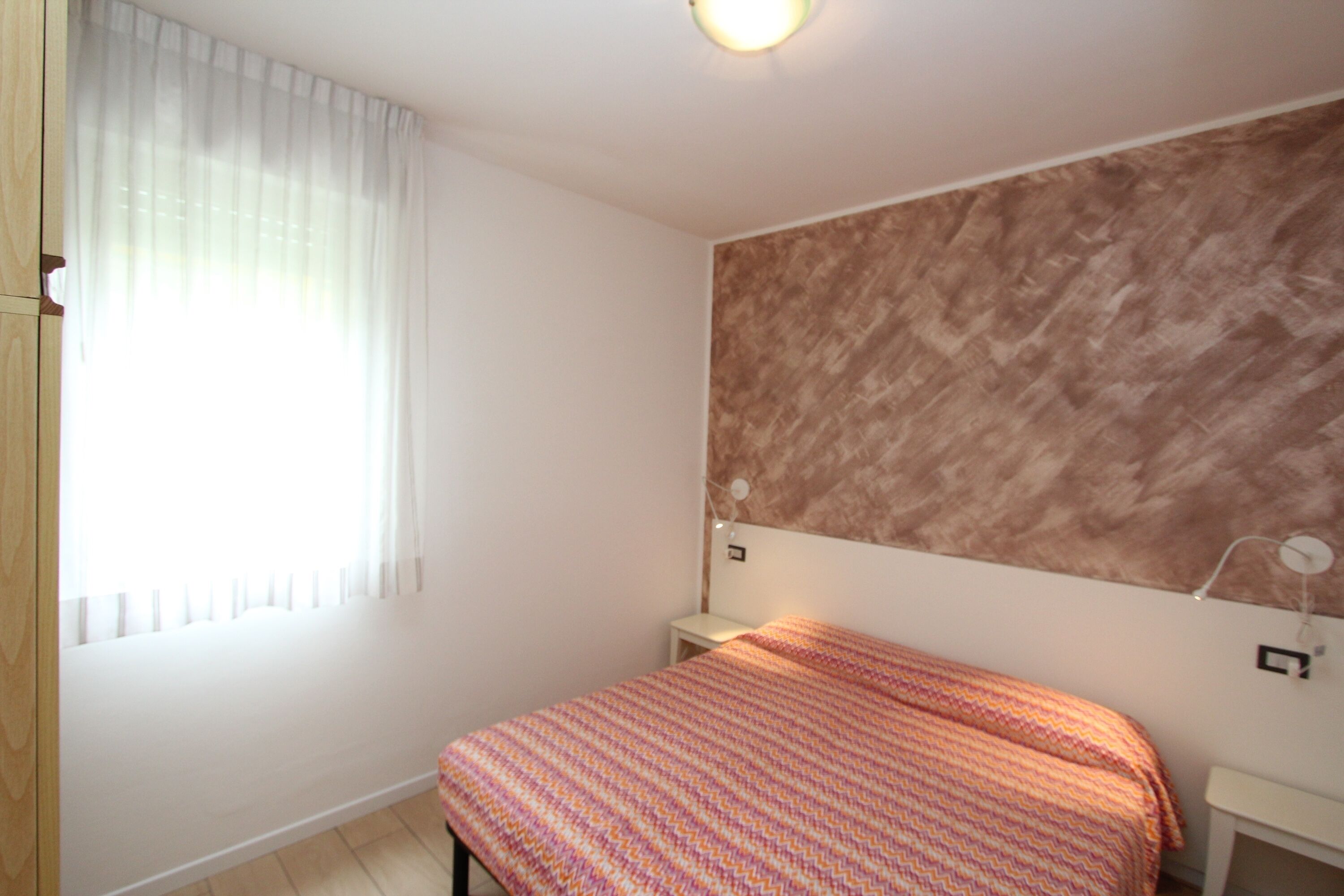 Un dormitorio con una cama, una mesita de noche, una lámpara de pared y una ventana con cortinas.