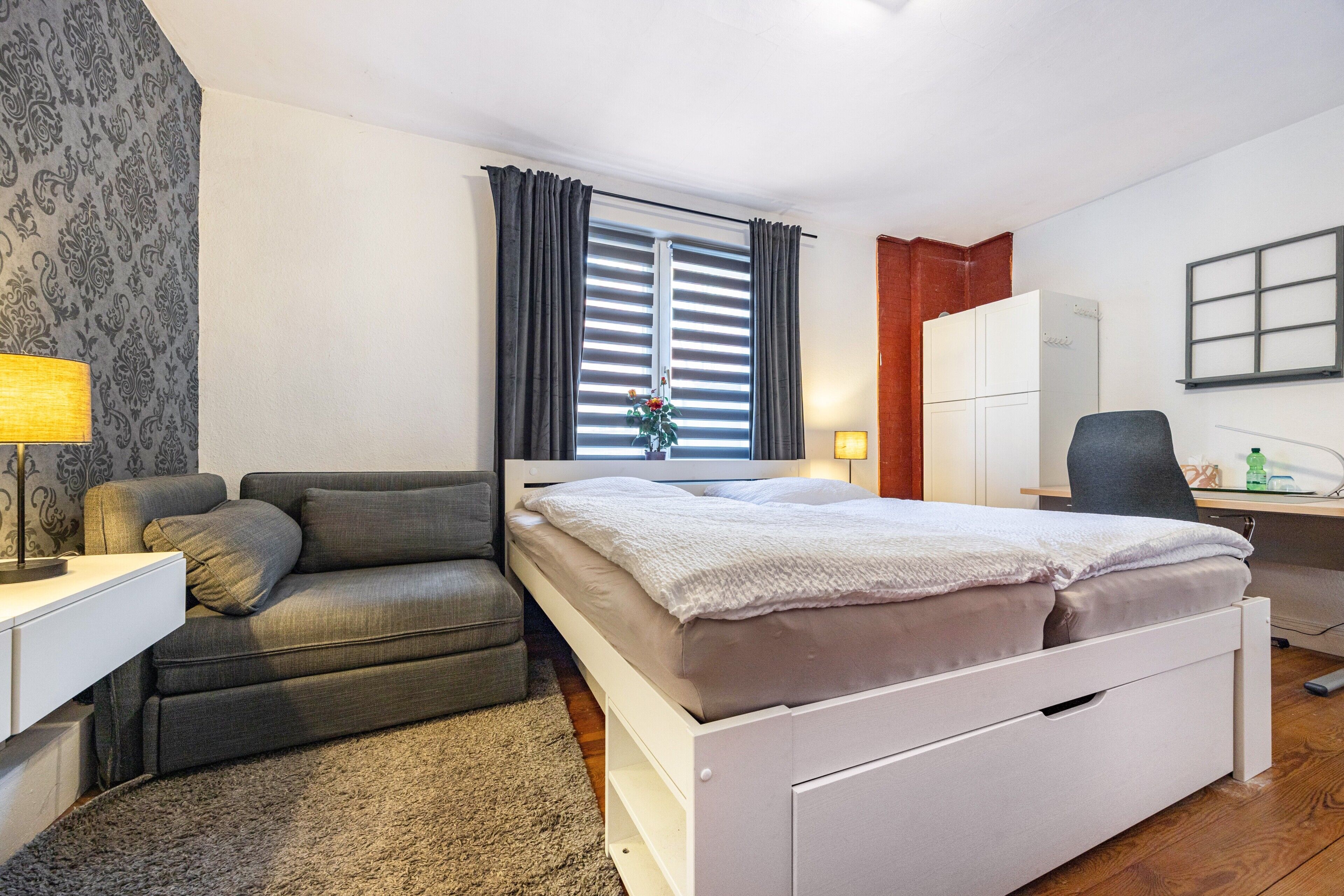 A bedroom with a bed, a desk, a chair, a wardrobe, and a window with curtains.
