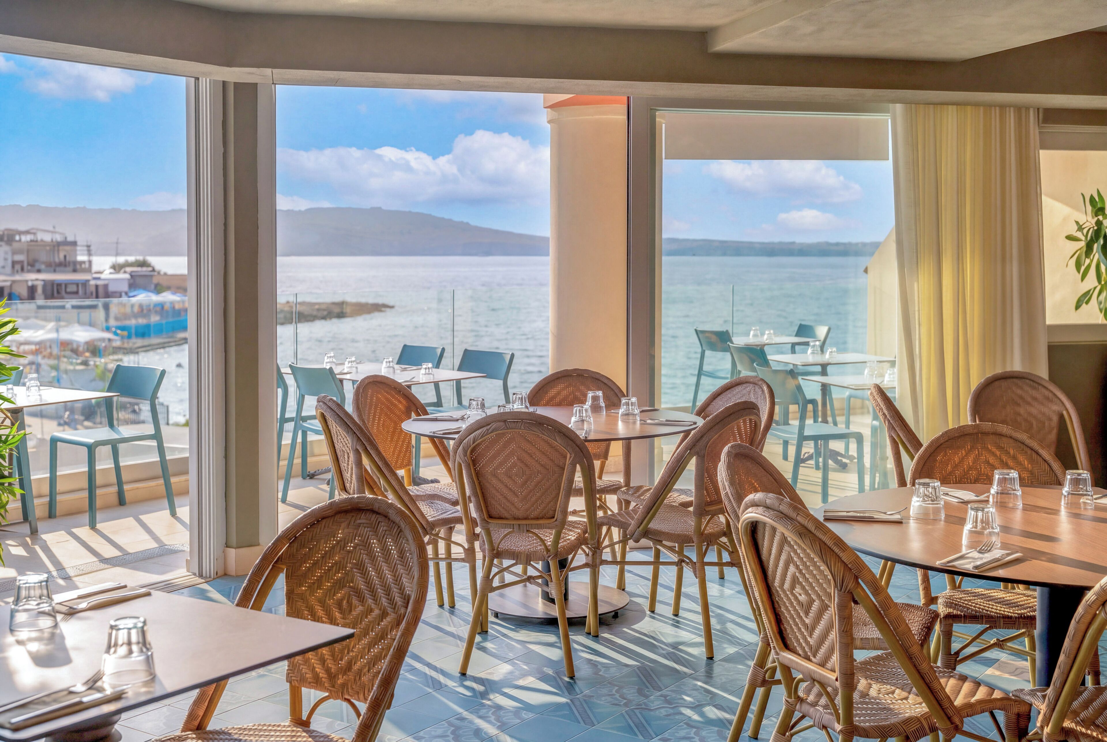 Un espace repas avec des chaises et des tables en osier, donnant sur une étendue d’eau dont on aperçoit la rive au loin.