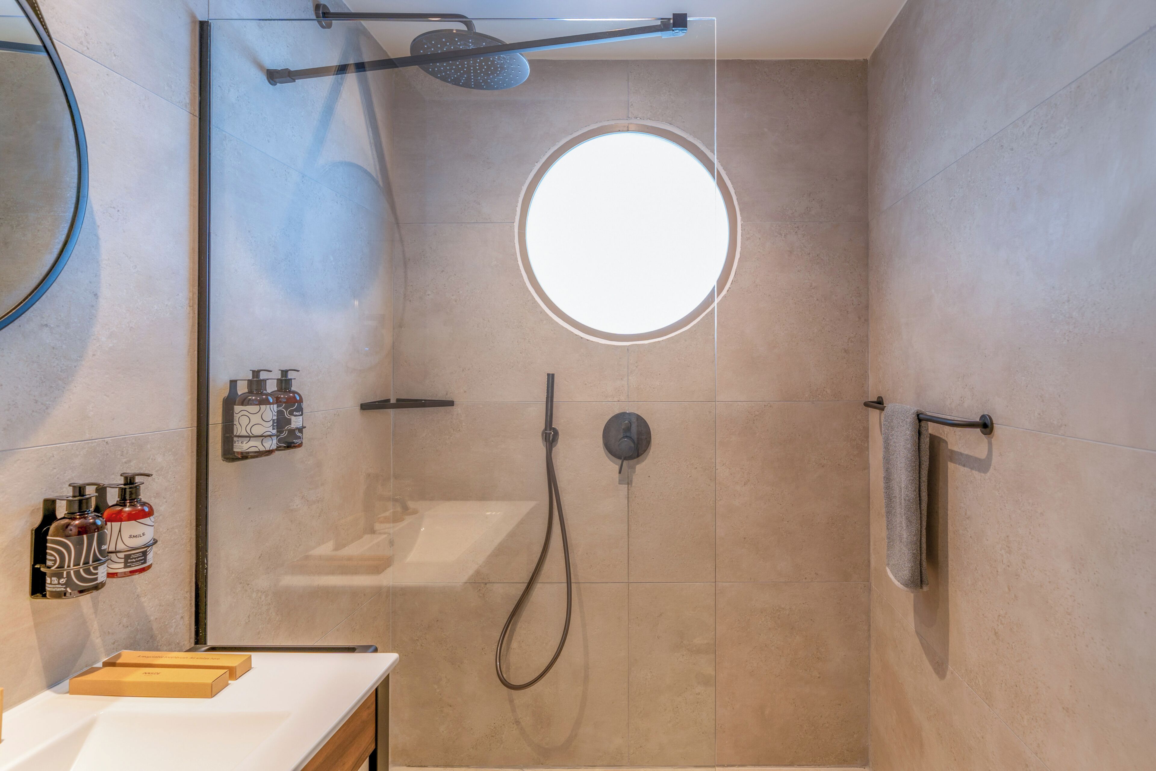 Un baño moderno con ducha envuelta en vidrio, un tragaluz circular y encimera blanca con artículos de aseo.