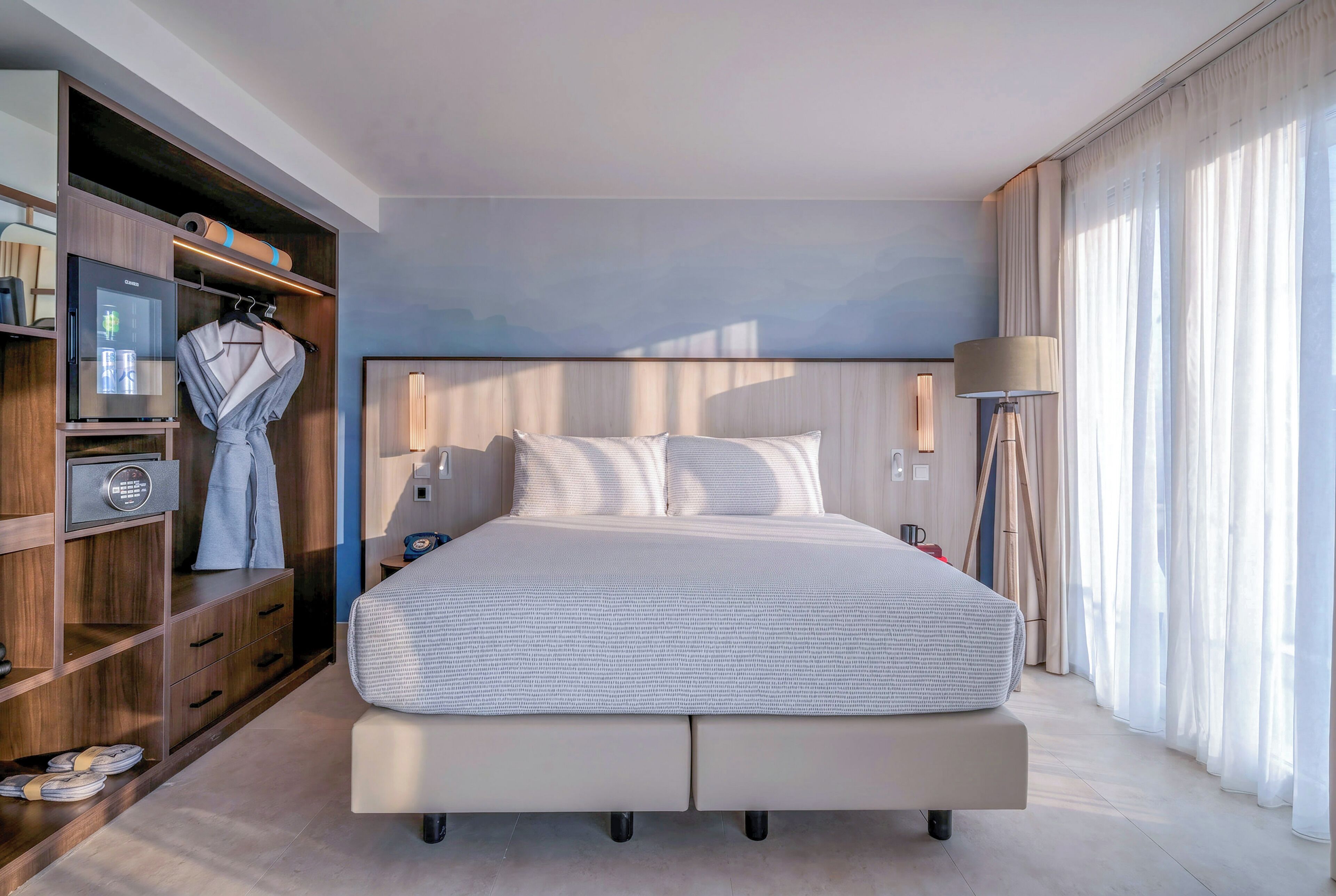 Une chambre d’hôtel moderne équipée d’un grand lit, d’une armoire, d’un mini-bar et d’une lampe de chevet.
