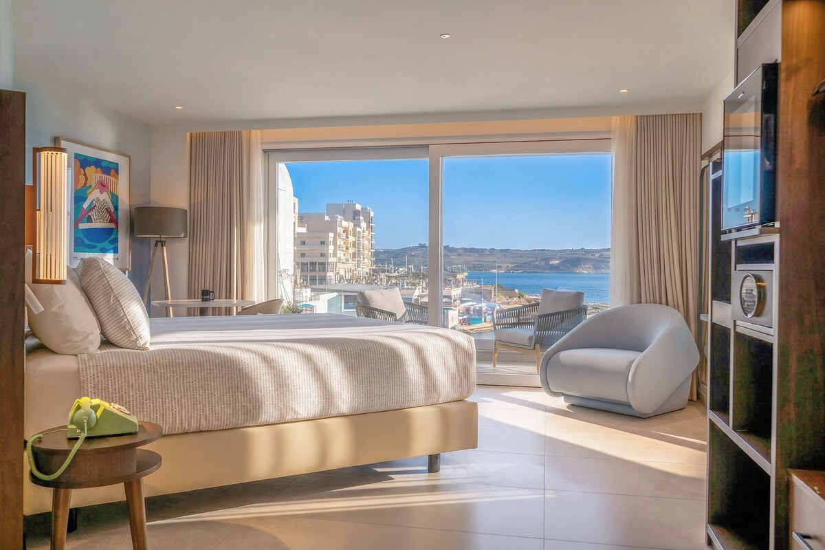 A modern hotel room with a large bed, a seating area with a chair, a small table, and a view of the city and sea.
