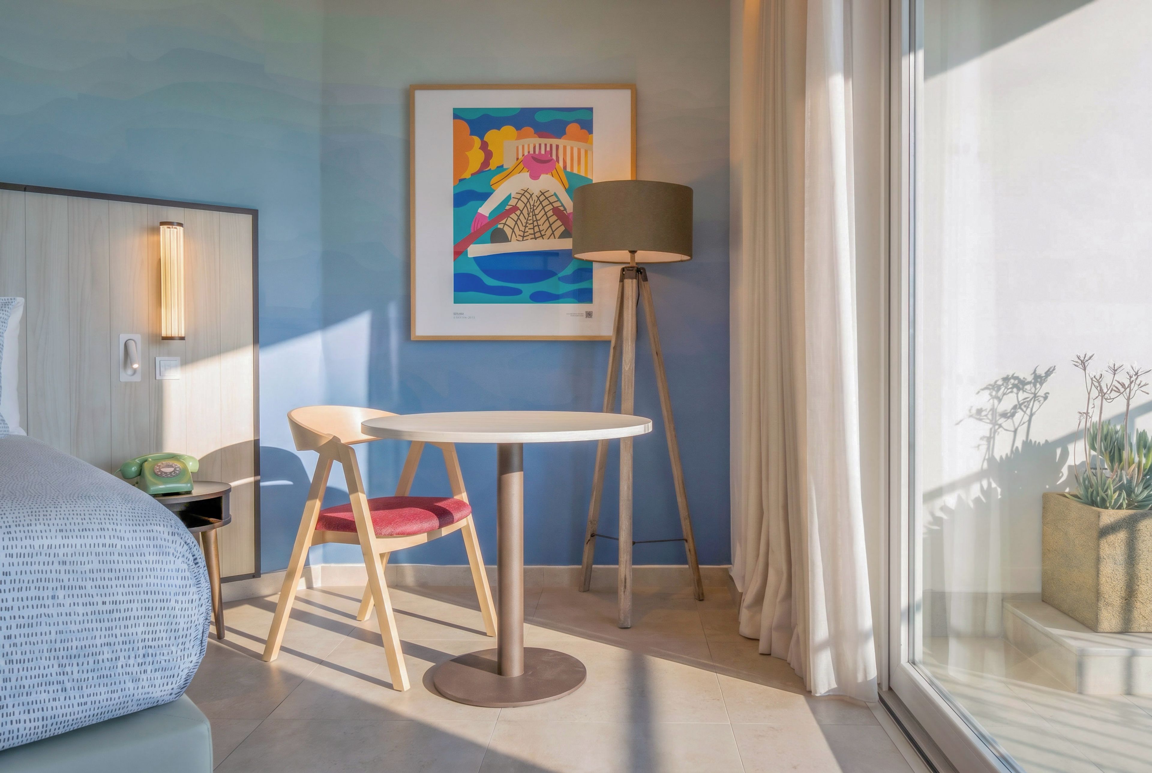 Habitación de hotel con una cama, una mesita con una silla, una lámpara de pie, un cuadro en la pared y una ventana con cortinas.