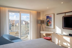 Habitación de hotel con cama, escritorio, silla, televisor, un cuadro y vistas a la ciudad.