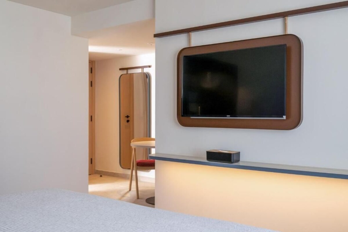 A modern hotel room with a large flat-screen TV mounted on the wall, a bedside table with a lamp, and a visible door leading to another room.