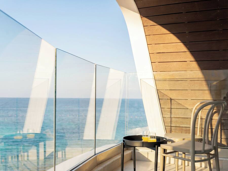 A balcony with a glass railing overlooking the sea, a small table with two glasses, and a chair.