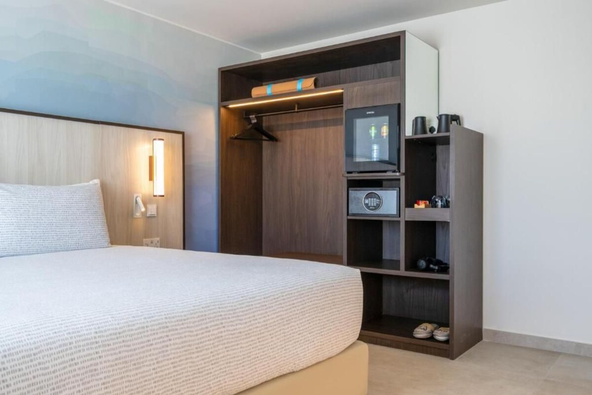 A hotel room with a bed, a wardrobe with shelves, a mirror, and a small table with a lamp.
