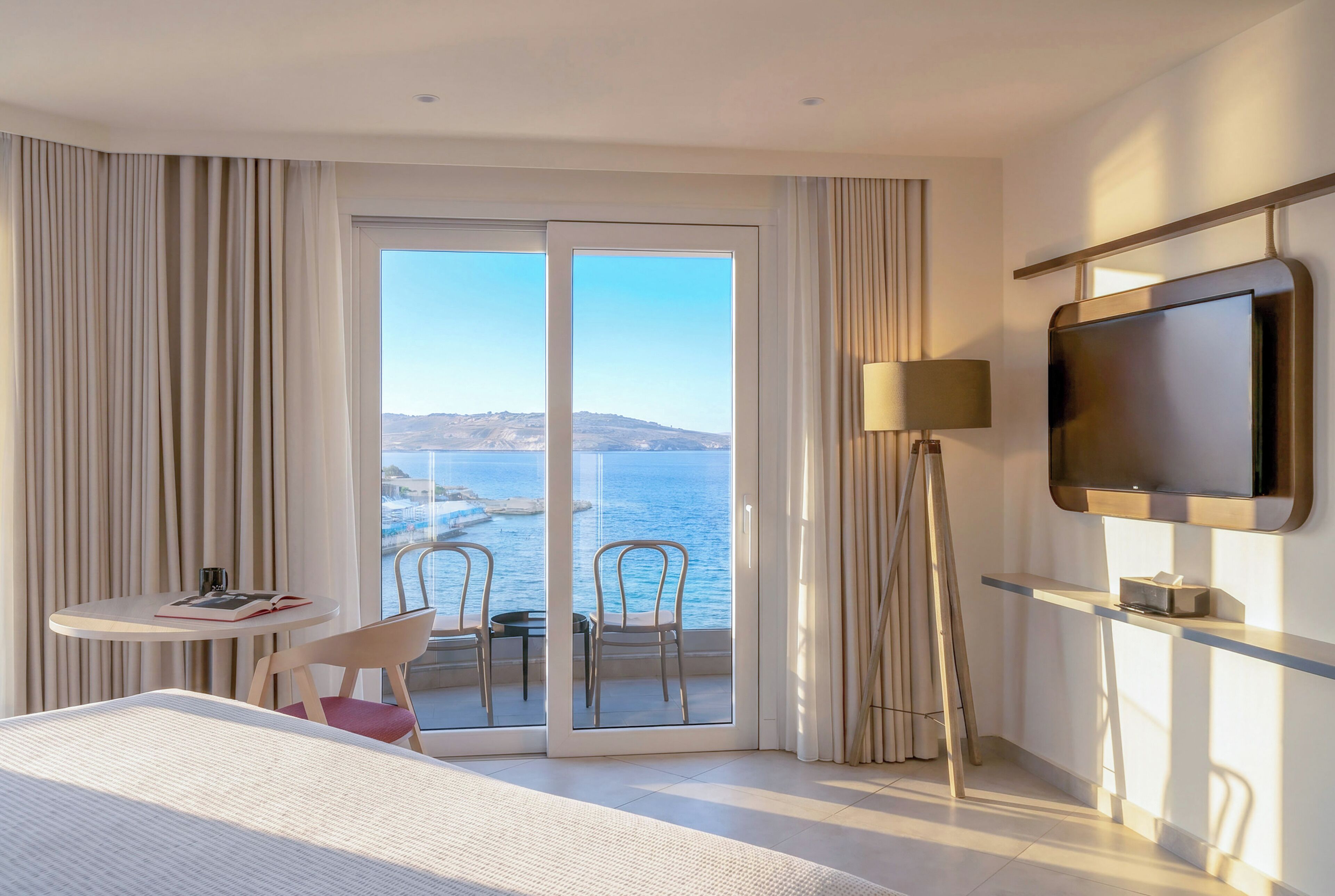 Habitación de hotel con cama, una pequeña mesa con silla, un televisor en la pared y balcón con vista al mar.