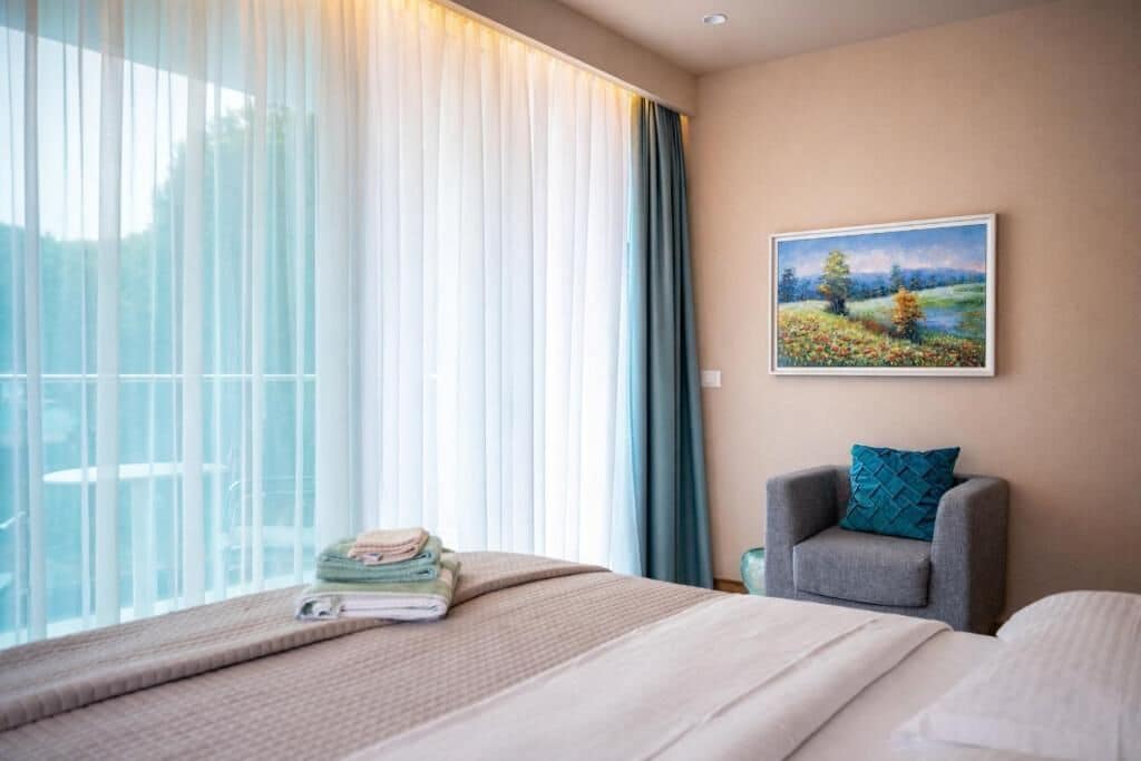 Une chambre d’hôtel avec un lit, une chaise, un tableau au mur et une fenêtre avec des rideaux.