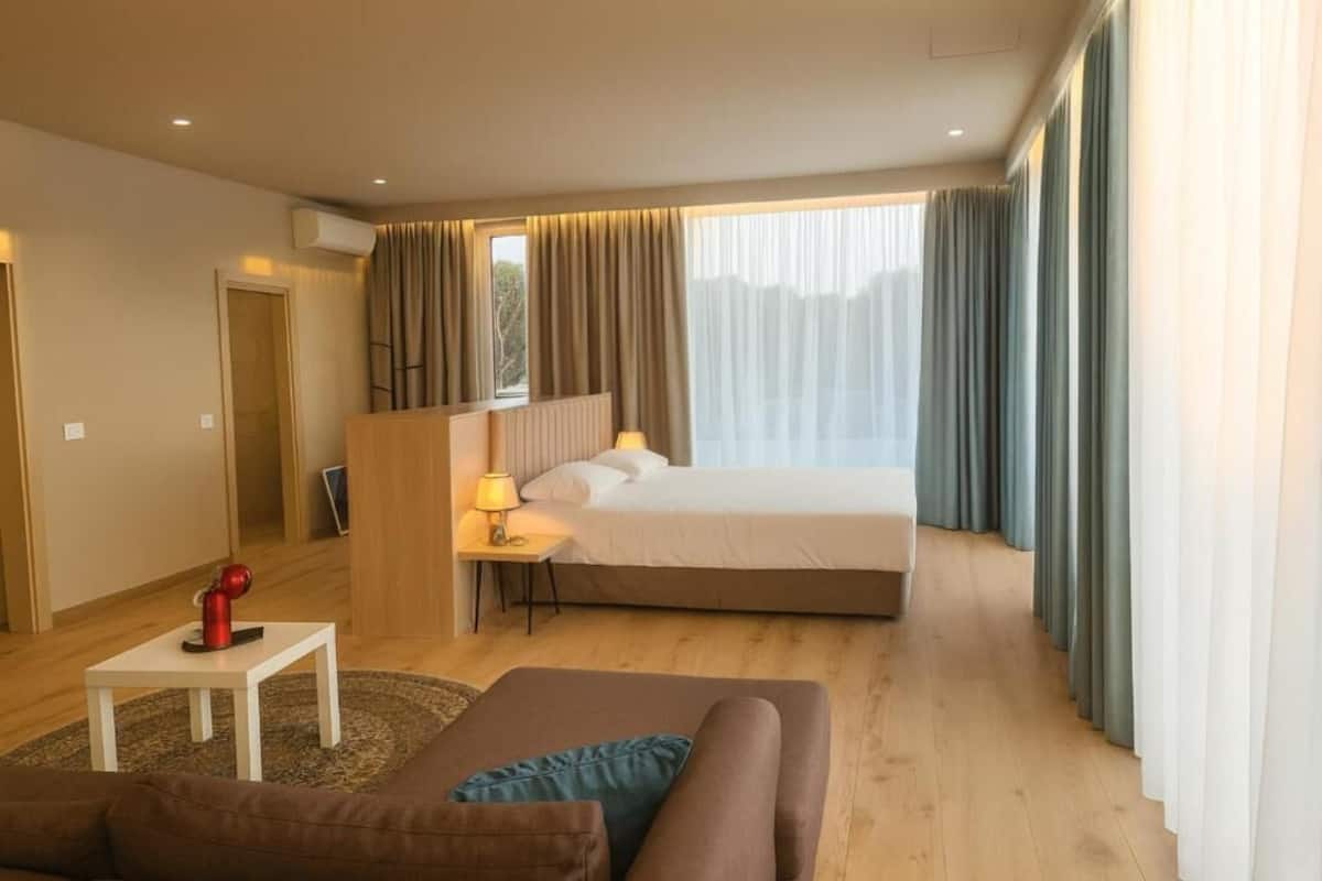 Una habitación de hotel con una cama, una mesita con una lámpara, un sofá y cortinas.