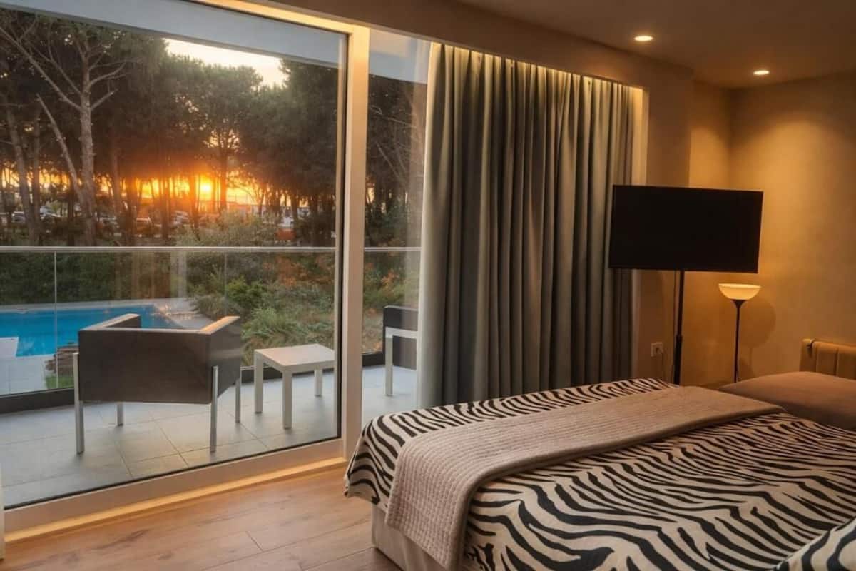 Un dormitorio con un ventanal grande, una cama con cubrecolcha de estampado de cebra, un televisor de pantalla plana y vistas a una piscina y árboles.