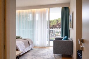 Una habitación de hotel moderna con una cama, un sillón gris y un ventanal con cortinas.