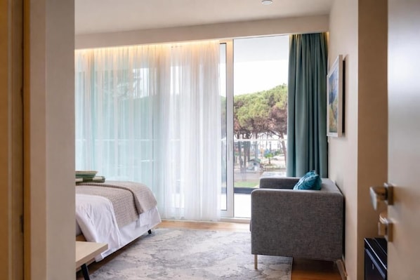 A modern hotel room with a bed, a grey armchair, and a large window with curtains. - Vila Eva (Golem)