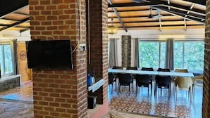 A spacious room with a brick fireplace, a dining area with a table and chairs, and large windows offering a view of the outdoors. - Gambhiraa Resorts (Kolli Hills)