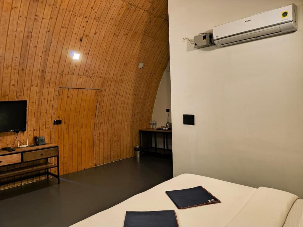 Een kamer met een gebogen houten plafond, een bed met een wit matras, een flatscreen televisie op een houten dressoir en een wandairconditioner.