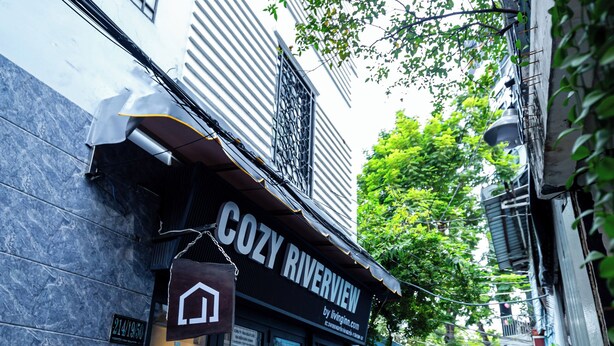En smal gränd med en byggnad som har skylten ”COZY RIVERVIEW”. Byggnaden har en modern fasad, en entré med glasdörr och ett litet utomhusområde med sittplatser.
