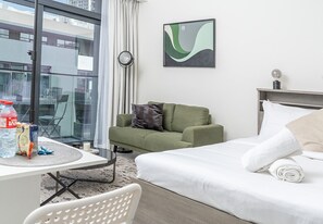 A modern hotel room with a bed, a green sofa, a dining table with a chair, a clock, and a view of the outside. - Modern Studio in Oxford Boulevard JVC (Dubai)